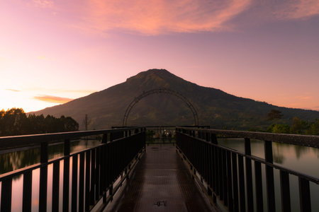 Mount Sumbing with sunrise sky and bridge on the foreground. There is artificial lake surrounded the bridge. Embung Kledung, Central Java, Indonesiaの写真素材