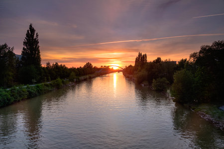 As the sun sets, its warm glow reflects on the tranquil water of the river, creating a picturesque scene surrounded by trees and a colorful afterglow in the skyの写真素材
