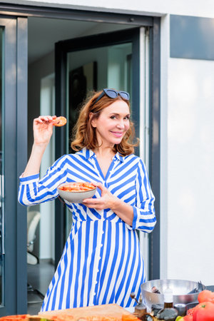Woman with brown hair in a blue and white striped dress prepares shrimp on a cozy outdoor patio with vegetables and natural light, creating a relaxed, inviting atmosphere.の写真素材