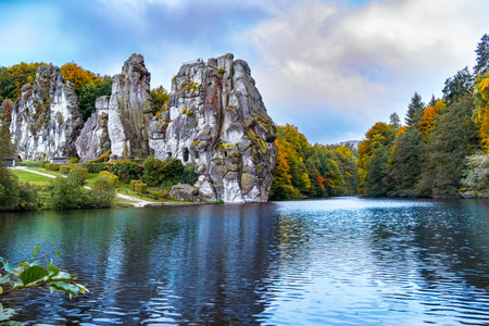 The Externsteine rock columns, standing 40 meters tall, form a striking natural monument in a peaceful nature reserve in Germany. Surrounded by autumnal colors, the reflection of the rocks and foliage create a picturesque scene on the lake.の写真素材