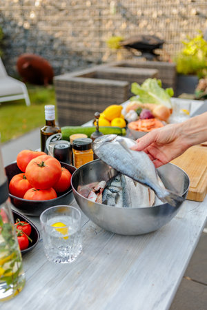Outdoor setup with fresh ingredients on a table, including whole fish, tomatoes, green peppers, and spices. A cozy patio setting with a natural, rustic atmosphere.の写真素材