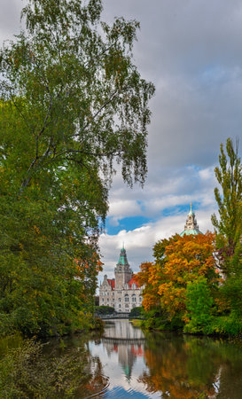 Colorful autumn view of the New Town Hall in Hannover, Germany, reflecting in the water of the surrounding park lake. Historic architecture framed by vivid trees and calm ripples.の写真素材