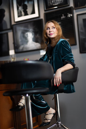 Elegant middle-aged woman in teal velvet suit sitting at bar counter with coffee and champagne. Background with framed black and white photographs. Cozy stylish atmosphere.の写真素材