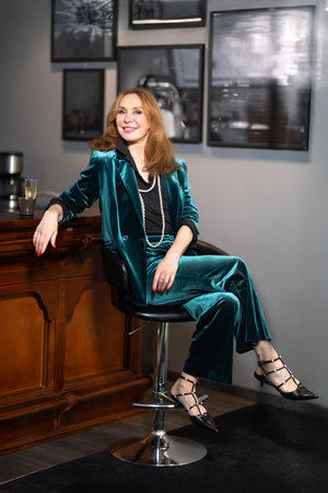 Elegant middle-aged woman in teal velvet suit sitting at bar counter with coffee and champagne. Background with framed black and white photographs. Cozy stylish atmosphere.の写真素材