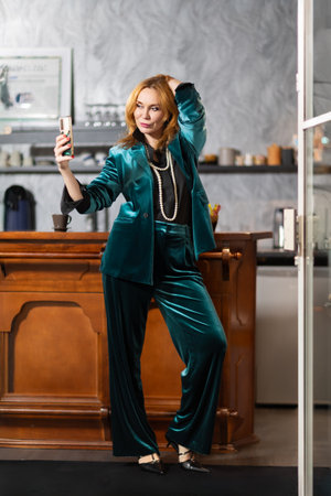 Elegant middle-aged woman in teal velvet suit sitting at bar counter with coffee and champagne. Background with framed black and white photographs. Cozy stylish atmosphere.の写真素材