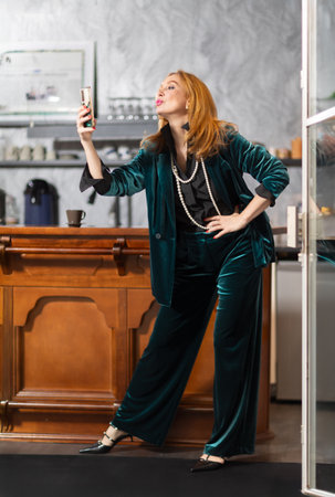 Elegant middle-aged woman in teal velvet suit at bar counter with coffee and champagne. Background with framed black and white photographs. Cozy stylish atmosphere.の写真素材