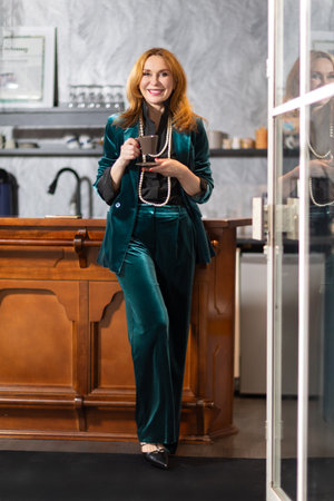 Elegant middle-aged woman in teal velvet suit sitting at bar counter with coffee and champagne. Background with framed black and white photographs. Cozy stylish atmosphere.の写真素材