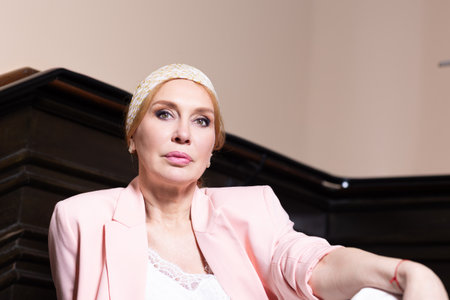 Elegant mature woman in a pastel pink blazer and white outfit posing on vintage staircase. Confident facial expressions and stylish outfit. Studio light indoor fashion portrait.の写真素材