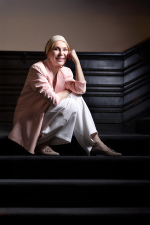Elegant mature woman in a pastel pink blazer and white outfit posing on vintage staircase. Confident facial expressions and stylish outfit. Studio light indoor fashion portrait.の写真素材