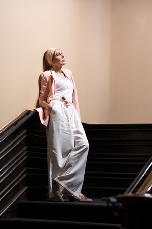 Elegant mature woman in a pastel pink blazer and white outfit posing on vintage staircase. Confident facial expressions and stylish outfit. Studio light indoor fashion portrait.の写真素材