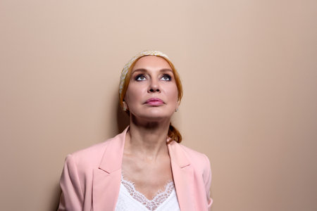 Elegant mature woman in a pastel pink blazer and white outfit posing on vintage staircase. Confident facial expressions and stylish outfit. Studio light indoor fashion portrait.の写真素材
