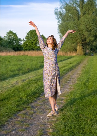 A cheerful woman in a long floral dress is enjoying nature, walking and dancing on a green path in the park. She looks free and happy, surrounded by trees and soft spring sunlight.の写真素材