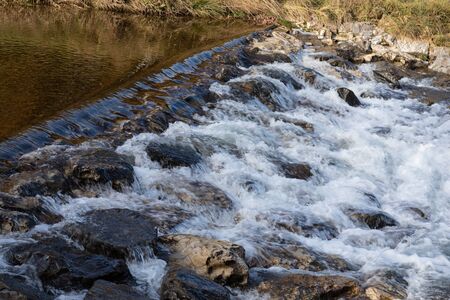 autumn waterfall with many large stones and meadow banks in the raging water side viewの写真素材