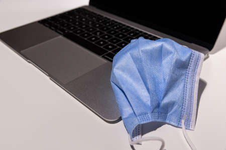 blue covid 19 mask on an opened silver laptop, image is strongly focused on the protective mask,...