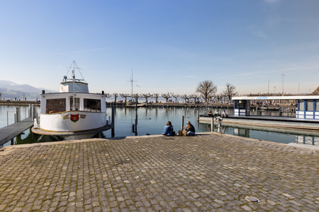 rapperswil jona, sankt gallen, switzerland - february 20, 2021: beach promenade, empty place with parked boat, two tourists sitting having lunch, in the background crippled trees without leaves, by dayのeditorial素材