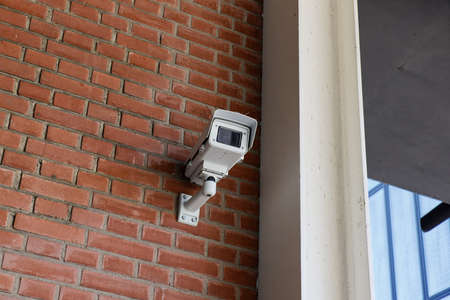 white surveillance camera mounted on a red stone wall, people are under constant surveillance in today's world, crime prevention, daytimeの写真素材