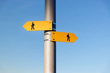 two yellow hiking trail signs with symbol person, backpack, hiking sticks, the signs point in...