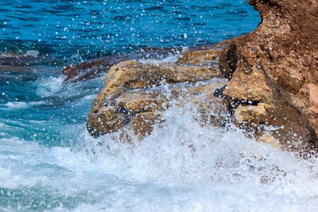Sea waves hit the rocky stones of coastline. Water surf with summer sunlight and water drops with foam.の写真素材