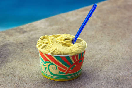 Scilla, Italy - September 7, 2019: Traditional italian gelato ice cream in paper cup on concrete wall of ancient town near the blue sea. Pistachio sweet cold dessert closeup outdoors.のeditorial素材