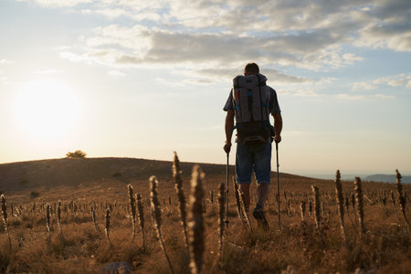 Young hiker with backpack and trekking poles walking along a mountain path at sunset with a smile. High quality photo.の写真素材