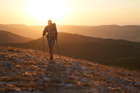 Young hiker with backpack and trekking crimea poles walking along a mountain path at sunset with a smile. High quality photo.の写真素材