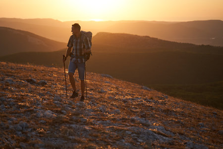 Young hiker with backpack and trekking poles walking along a mountain path at sunset with a smile. High quality photo.の写真素材