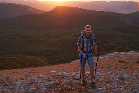 Young hiker with backpack and trekking poles walking along a mountain path at sunset with a smile. High quality photo.の写真素材