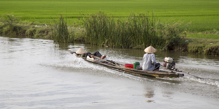 Hue, Vietnam - Jun 17, 2016: Vietnamese farmer with traditional conical hat driving a machine wooden boat on a canal providing water for the green paddy field.のeditorial素材