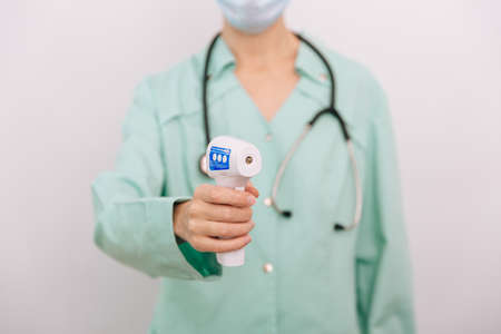 Temperature measurement gun in doctor hands. Close-up shot of doctor wearing protective surgical mask ready to use infrared isometric thermometer gun to check body temperature for virus symptoms.の写真素材