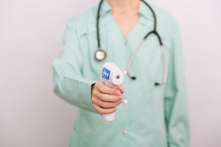 Temperature measurement gun in doctor hands. Close-up shot of doctor wearing protective surgical mask ready to use infrared isometric thermometer gun to check body temperature for virus symptoms.の写真素材