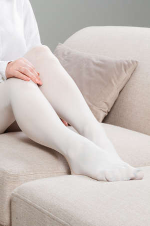 White hosiery. Beautiful long female legs in stockings. Girl putting on stockings at home in a white room. Varicose veins prevention. Woman body in underwear.の写真素材