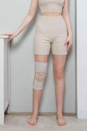 Knee support brace on a woman leg. girl in an orthosis in the interior of the house. Orthopedic anatomic braces for knee fixation, injuries and pain. Knee Joint Bandage Sleeve. Elastic Sports.の写真素材