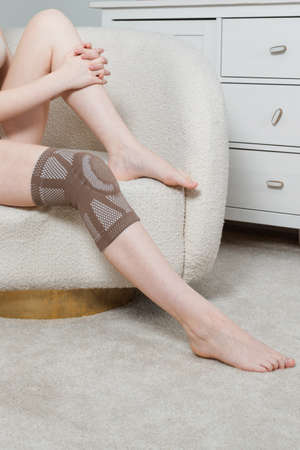 Knee support brace on a woman leg. girl in an orthosis in the interior of the house. Orthopedic anatomic braces for knee fixation, injuries and pain. Knee Joint Bandage Sleeve. Elastic Sports.の写真素材