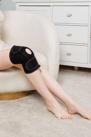Knee support brace on a woman leg. girl in an orthosis in the interior of the house. Orthopedic anatomic braces for knee fixation, injuries and pain. Knee Joint Bandage Sleeve. Elastic Sports.の写真素材