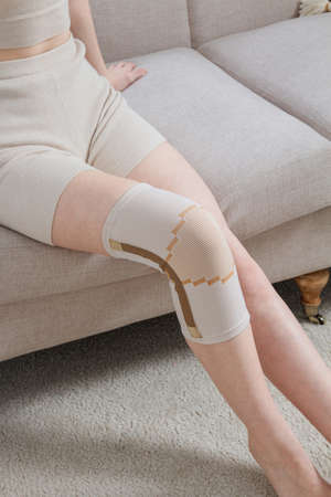 Knee support brace on a woman leg. girl in an orthosis in the interior of the house. Orthopedic anatomic braces for knee fixation, injuries and pain. Knee Joint Bandage Sleeve. Elastic Sports.の写真素材