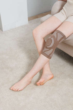 Knee support brace on a woman leg. girl in an orthosis in the interior of the house. Orthopedic anatomic braces for knee fixation, injuries and pain. Knee Joint Bandage Sleeve. Elastic Sports.の写真素材