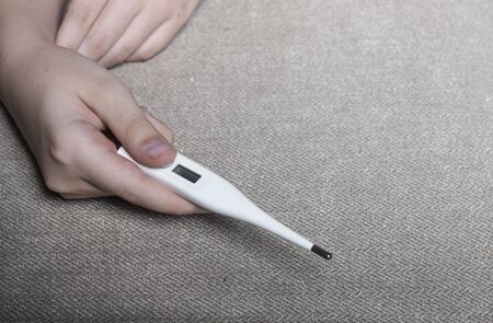 The hand of a child who holds out into the frame a modern electronic thermometer in white. Background is a rough textured gray fabric.の写真素材