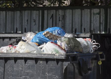 A garbage container. A large black container filled with debris. Bags of garbage of different colors and a mountain rise above the container. There 's a fence in the background. Sunny Morningの写真素材