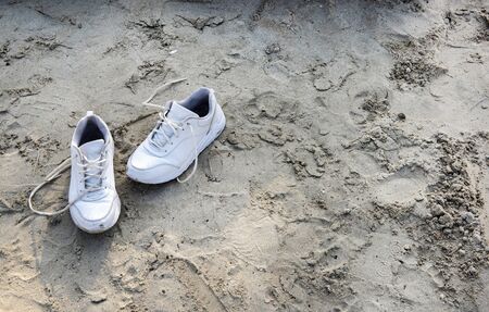 A beach, light dense sand on which stands a pair of white sneakers with untied white shoelaces. The morning light of the bright sunの写真素材