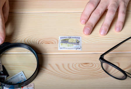 Moscow, Russia. 02-24-2020. In the center of the wooden table is a postage stamp with a stamp on which the bridge is depicted. Hands jo man are holding a magnifying glass, glasses are nearby.のeditorial素材
