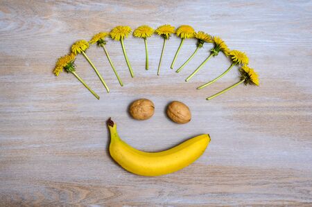 A comic image of a smiling face on a light wooden surface. Banana is a smile, walnuts are eyes and dandelions are like a short hairstyle.の写真素材