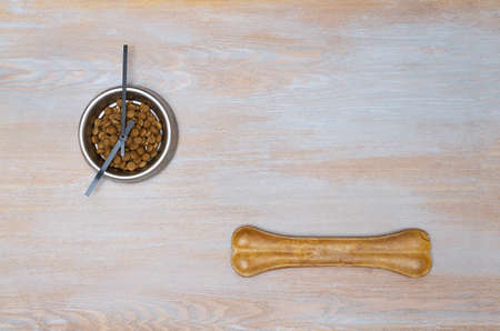 Meal time is dog lunch. A bowl of dry food for dogs on which the clock hands and a large bone lie. A vintage wooden surface with a bowl of food and a large bone on it.の写真素材