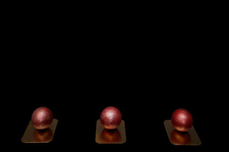 Three festive Easter eggs painted with red paint lie in one line on gold coasters against a large black background - top view. Studio photography on a large banner.の写真素材