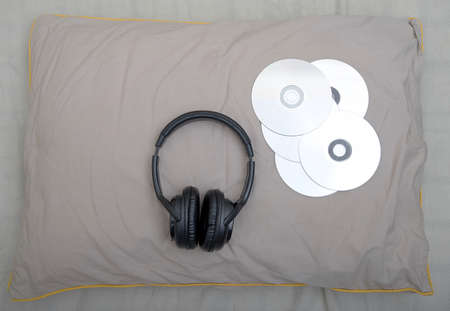 Large stylish black headphones for listening to music and several CDs lie on a soft gray pillow in the bedroom. View from above.の写真素材