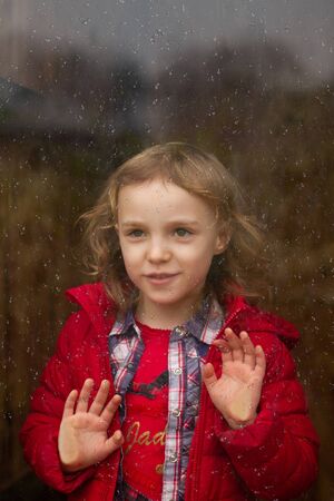 beautiful girl in a red jacket looks through the glass windowの写真素材