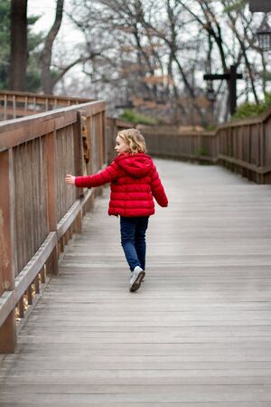 A little girl in a red jacket walks down the road touching the fenceの写真素材