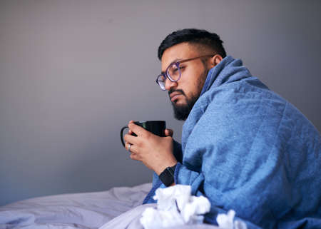 A sick man sits in bed wrapped in a blanket holding a warm drink. Copy spaceの写真素材