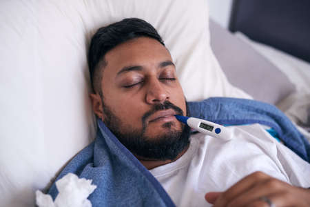 A sick man lies with an oral thermometer reading 38.3 degrees Celcius for feverの写真素材