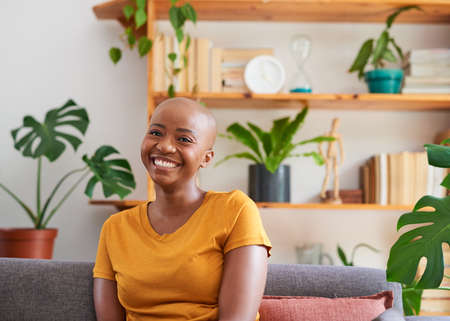 A young woman smiles while sitting on the couch- bookshelf filled with plants