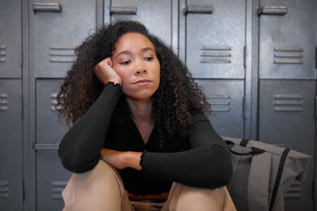 A young mixed race woman sits in front of school lockers looking depressed